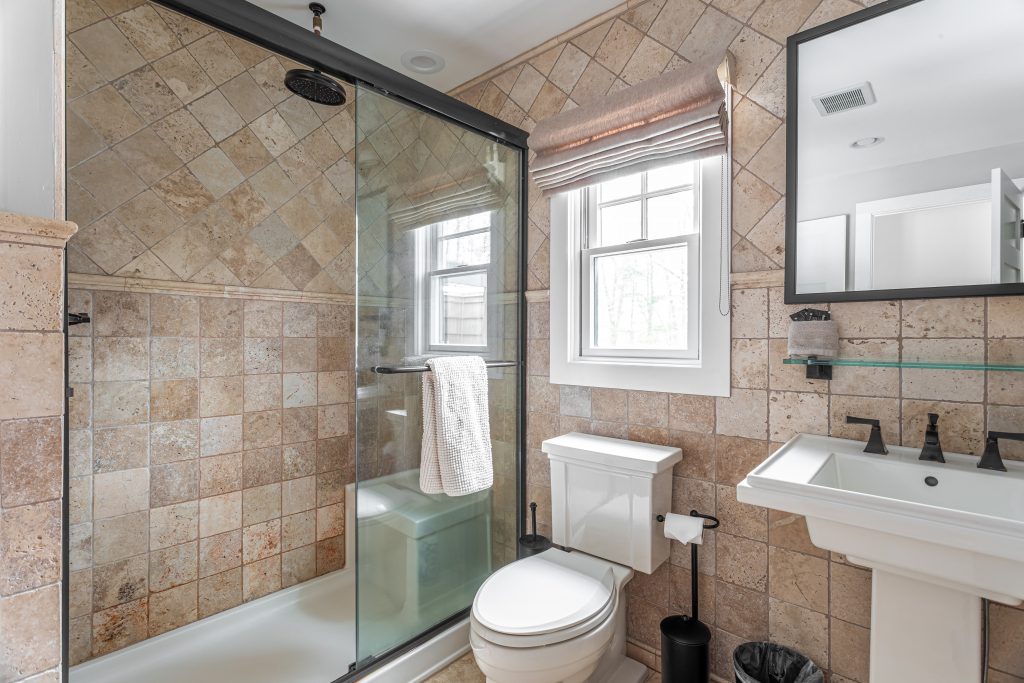 Bathroom with tiled walls, glass shower, pedestal sink, toilet, and window with natural light. Warm, classic interior design with neutral stone textures.